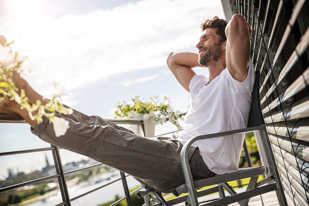 Männliches Model sitzt auf einem Stuhl auf dem Balkon mit den Beinen auf dem Geländer